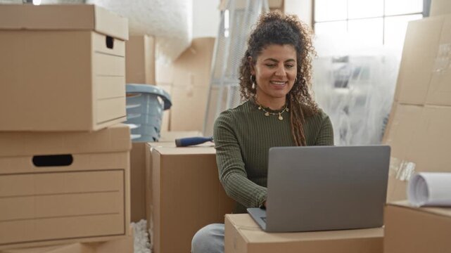 Woman smiling and typing on a laptop amid stacked moving boxes while seated in a building; moving optimism.