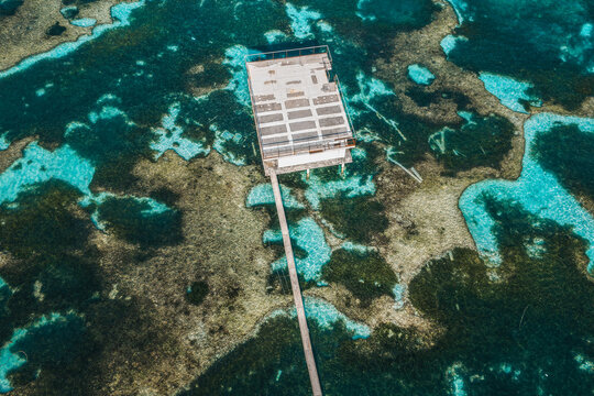 Aerial view of a modern overwater structure and wooden pier surrounded by vibrant coral reefs and turquoise ocean water in Kudahuvadhoo, Dhaalu Atoll, Maldives.