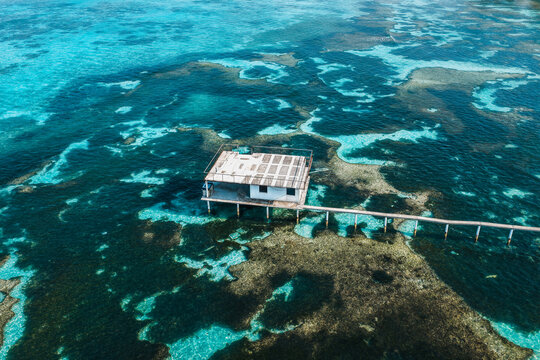 Aerial view of a small white building on stilts connected by a long wooden pier over a vibrant coral reef in turquoise waters in Kudahuvadhoo, Dhaalu Atoll, Maldives.