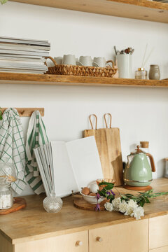 Close-up of a kitchen counter and shelves with assorted items including an open cookbook, kettle, fresh eggs, chopping boards, flowers, tea towels and crockery