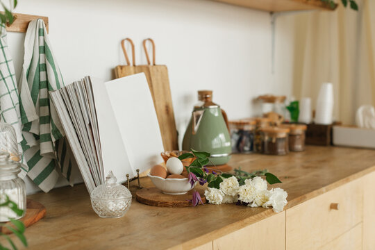 Close-up of a kitchen counter with assorted items including an open cookbook, kettle, fresh eggs, chopping boards, flowers, tea towels and pots