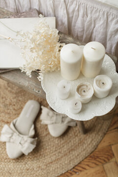 Close-up overhead view of assorted block candles and dried flowers on a wooden bench at the end of a bed with a pair of slippers and glass vase