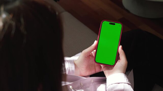 Over shoulder view of woman holding smartphone with green screen mockup at home. Mobile phone display ready for screen replacement, app presentation, advertising or digital content. High quality 4k