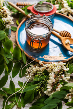 Close-up of a glass Jar of spring honey with a honey dipper and tag with the word honey on a table with fresh Acacia flowers