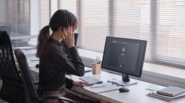 Medium long shot of young female Asian customer service specialist using headset with microphone and calling on computer at desk in office
