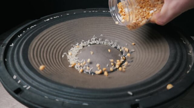 White rice grains dance on a large black speaker cone while a hand pours yellow split peas onto the vibrating surface. Concept of acoustics, sound physics, and frequency visualization.