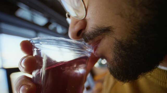 Close up tracking shot of young bearded man drinking from plastic cup in modern fast food restaurant