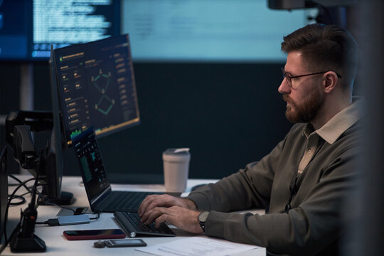 Caucasian young adult man analyzing cybersecurity data on multiple screens and typing on laptop in office. Side view, ideal for IT security, data monitoring, risk management marketing