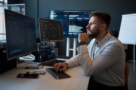 Caucasian young adult man analyzing cybersecurity data on multiple monitors in office, monitoring code and dashboards. Ideal for IT security, data analytics, tech workplace marketing