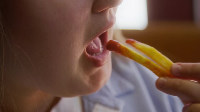Extreme close up shot of young woman eating french fries, dipped in ketchup while eating at restaurant