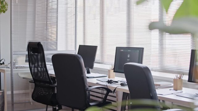 Establishing shot of computers monitors on tables, chairs and headsets in modern call center office, no people