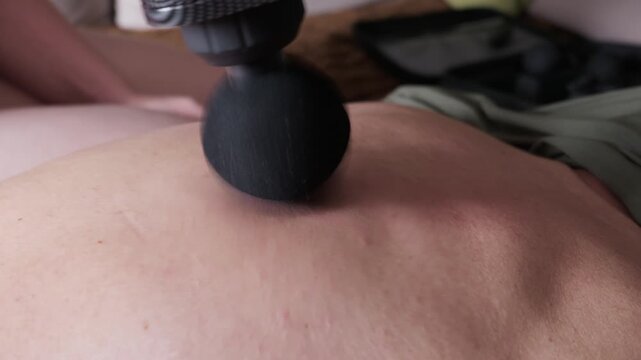 A black percussion massager vibrates deeply into bare back muscles. Concept of physical therapy and athletic recovery in an authentic home setting.