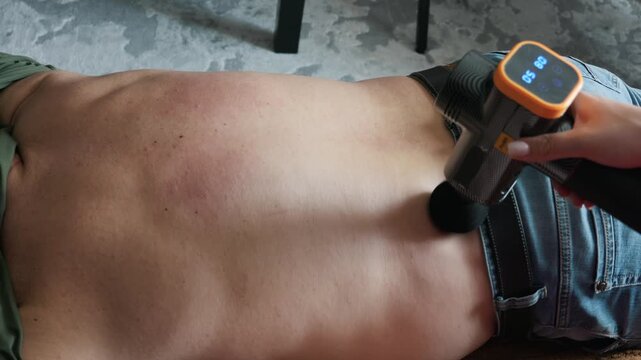 Partner uses a modern percussion massage gun on a man bare lower back to relieve muscle tension at home. Concept of physical therapy, pain relief and domestic athletic recovery.