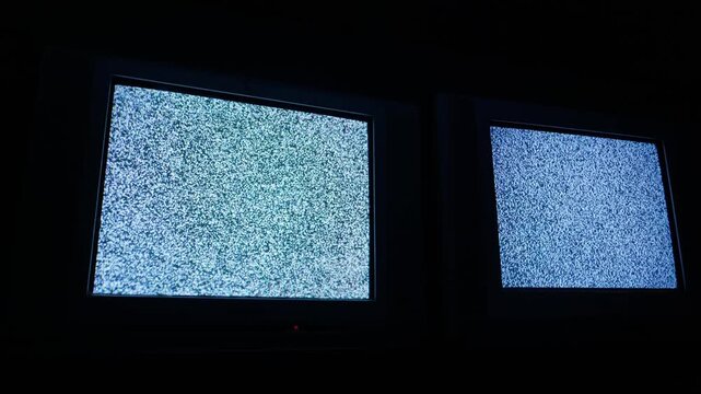 Two old cathode ray tube televisions flicker with white noise static in a pitch-black room, casting a dim blue glow. Concept of eerie technical failure and 1990s nostalgia.