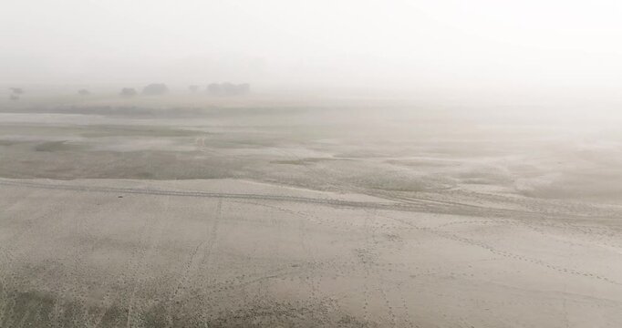 Aerial view of a vast sandy riverbed covered in thick fog with distant trees and footprints on the ground in Kurigram, Rangpur Division, Bangladesh.