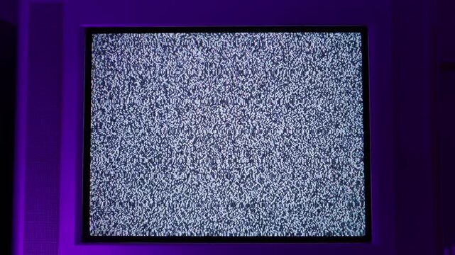 Vintage CRT television set displays white noise and signal interference on the screen. The monitor is surrounded by purple ambient lighting in a dark room. Concept of signal loss.