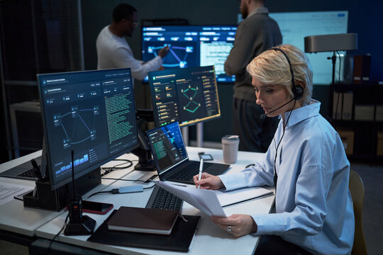 Caucasian middle aged woman wearing headset, reviewing reports and monitoring cybersecurity dashboard at workstation while colleagues analyzing data. For IT operations, network security, SOC marketing