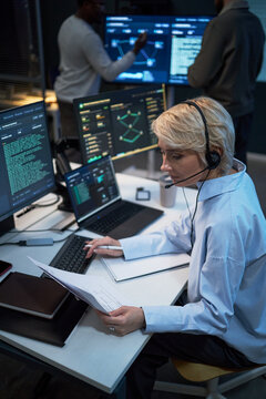Vertical shot of middle aged Caucasian woman monitoring cybersecurity data, reviewing report, using headset at control desk. Ideal for IT operations, network security, tech support