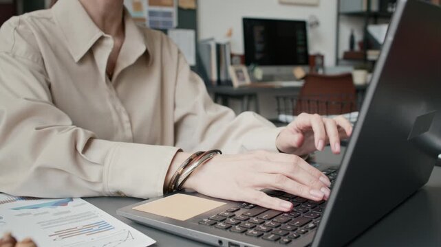 Low angle tilt up close up shot of focused Caucasian woman in blouse typing on laptop in modern cluttered office