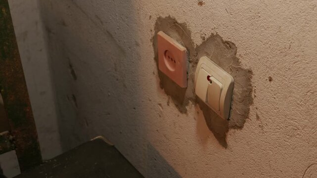A male hand reaches out in a dimly lit room to press a light switch mounted on a rough concrete wall. Concept of energy consumption, electrical power, and overcoming darkness.