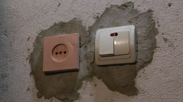 A hand presses a white light switch next to an electrical outlet on a freshly plastered wall. Concept of home renovation, interior construction, and DIY electrical repair.