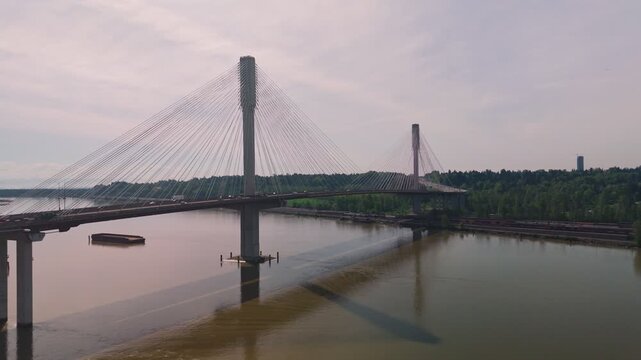 Port Mann Bridge cable architecture crossing wide calm murky river water carrying heavy transit past floating industrial barge casting long shadows over surface below hazy bright overcast sky reverse