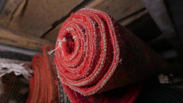 A close-up view shows the end of a thick hand-woven red rug rolled up tightly. Other patterned carpets and a rustic wooden beam are visible in the background. The wool textures are defined.