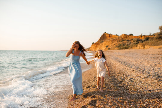 Happy woman with child girl daughter 5-6 year old holding hands walking at beach over sea at background. Summer vacation time. Motherhood.