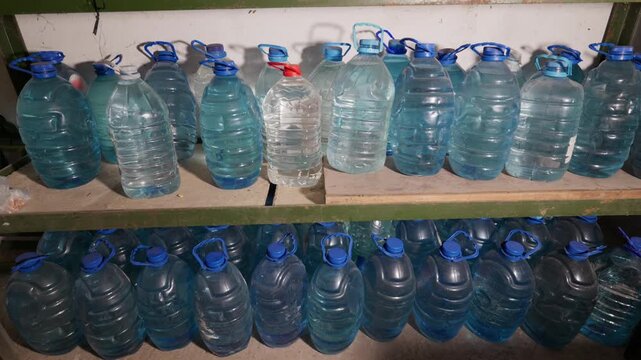 A large collection of five-liter plastic water jugs arranged neatly on wooden shelves in a storage room. Concept of emergency preparedness, survival supplies, and hydration planning.