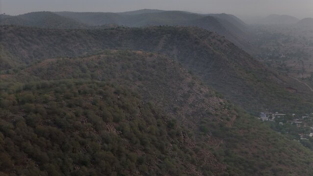View of rolling hills of the Aravalli Range covered in green shrubs under a hazy sky near the village of Naugaon, Rajasthan, India.