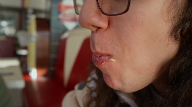 Side extreme close-up of face and mouth of young gen Z Caucasian man biting into tasty extra large hamburger and chewing, while enjoying visit to fast food restaurant with friends