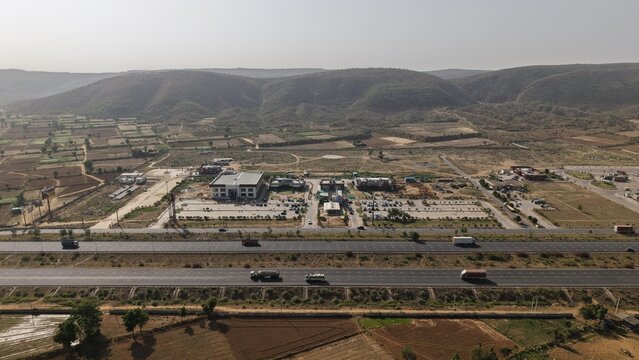 View of a modern highway rest area with trucks and cars parked near buildings against a backdrop of rolling hills in Naugaon, Rajasthan, India.