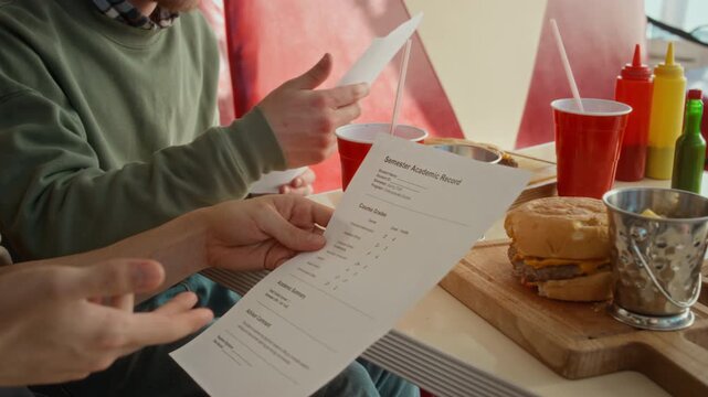Medium close-up of hands of unrecognizable young male college or university students checking out semester score records, comparing academic grades at cafe, burgers, chips and soda on table