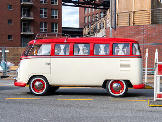 Obraz premium Classic red and white retro camper van parked beside modern brick buildings in an urban district, combining nostalgic road trip culture with vintage automotive design and travel lifestyle