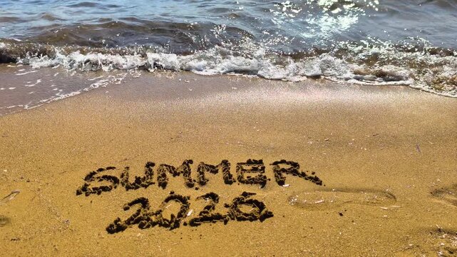 Summer 2026 text written on beach sand in slow motion on a Mediterranean beach at golden hour with warm cinematic light.