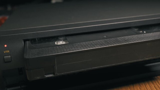 A black VHS cassette tape is slowly ejected from the slot of a vintage video player. The textured plastic casing reflects soft lighting, emphasizing the retro analog mechanics of the