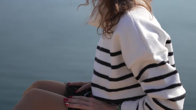 Woman, seaside, contemplation. Young female enjoying serene ocean view wearing striped sweater on a sunny day.