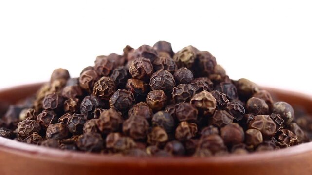Slowly rotating pile of whole black peppercorns in wooden brown bowl on white background, aromatic spice ingredient.
