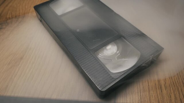 Black VHS cassette tape lies on a textured wooden table shrouded in thick white fog. Swirling mist slowly clears to reveal the retro analog media object under soft studio lighting.