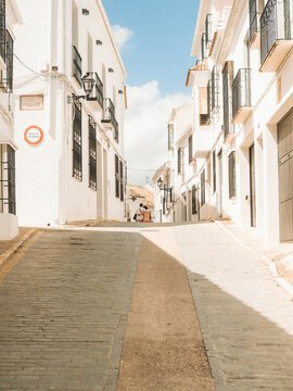 View of a narrow cobblestone street lined with traditional white-washed houses and two people walking in the distance under a bright blue sky in Zuheros, Andalusia, Spain.