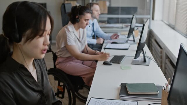 Slow motion handheld shot of Asian call center specialist coworking with female colleague in wheelchair and Caucasian man, using headsets and computers while providing remote support to customers