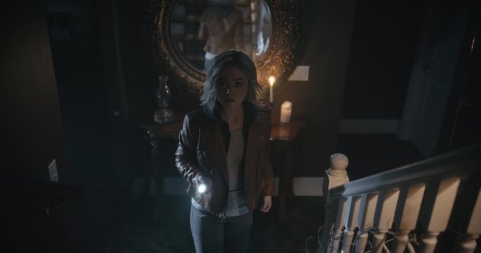 Woman staring at old mirror then going up spooky stairs with flashlight until something downstairs distracts her - high shot