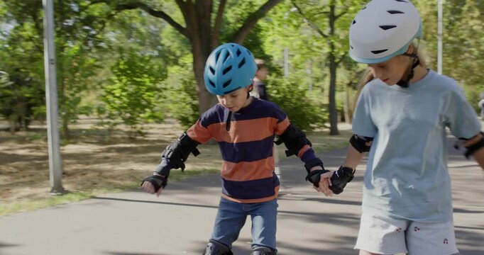 Brother and sister skate side by side while holding hands on park path. Children in helmets keep knees bent moving slowly together to maintain balance safely outside