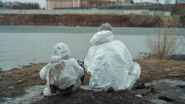 Caucasian homeless survivors by river, wrapped in white protective suits with muddy backs, facing industrial skyline across gray water, tense quiet vigil, hopeful glance toward distant shore and plans