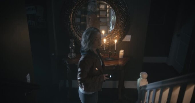 Woman with flashlight staring at reflection in large antique mirror then going upstairs until something distracts her - high, steady cam shot