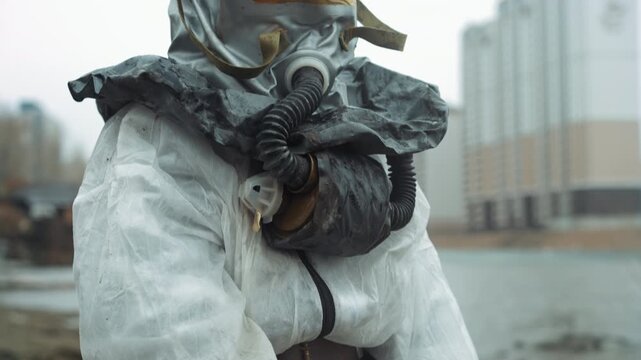 Scientist in protective suit exploring abandoned waterfront, closeup of stained coverall and respirator, blurred city skyline in background, cold overcast light, muddy shore and cautionary atmosphere