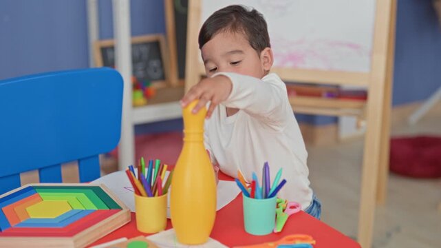 Boy reaching with hand for yellow toy bottle on a play table, holding pink crayon near colored pencils, wooden stacking puzzle and drawing paper in studio; curiosity learning discovery.