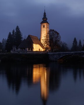 View of the illuminated Church of St. John the Baptist with its tall steeple reflected in the calm water of Lake Bohinj at dusk in Municipality of Bohinj, Radovljica, Slovenia.