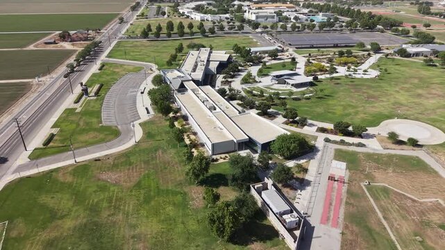 Drone view orbiting the modern architectural buildings of the College of the Sequoias campus, showcasing the surrounding green lawns, parking lots, and agricultural fields in Visalia, California