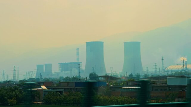 A moving side-view shot of a large industrial power plant near Xi'an, China. Massive cooling towers dominate the landscape, with smoke plumes rising against a hazy mountain silhouette.